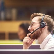 Businessman In Cubicle Wearing Headset Talking To Caller In Busy Customer Services Centre